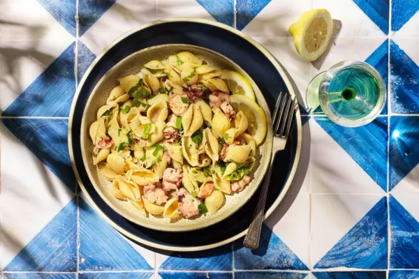 Un plat de pâtes aux coquillages et morceaux de poulpe, garni de persil frais et de quartiers de citron. Un verre de vin blanc et une fourchette complètent cette scène estivale.