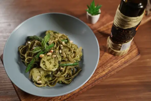 Assiette de trenette al pesto, garnie de haricots verts, pignons de pin et basilic frais. Une bouteille d'huile d'olive est visible à côté.